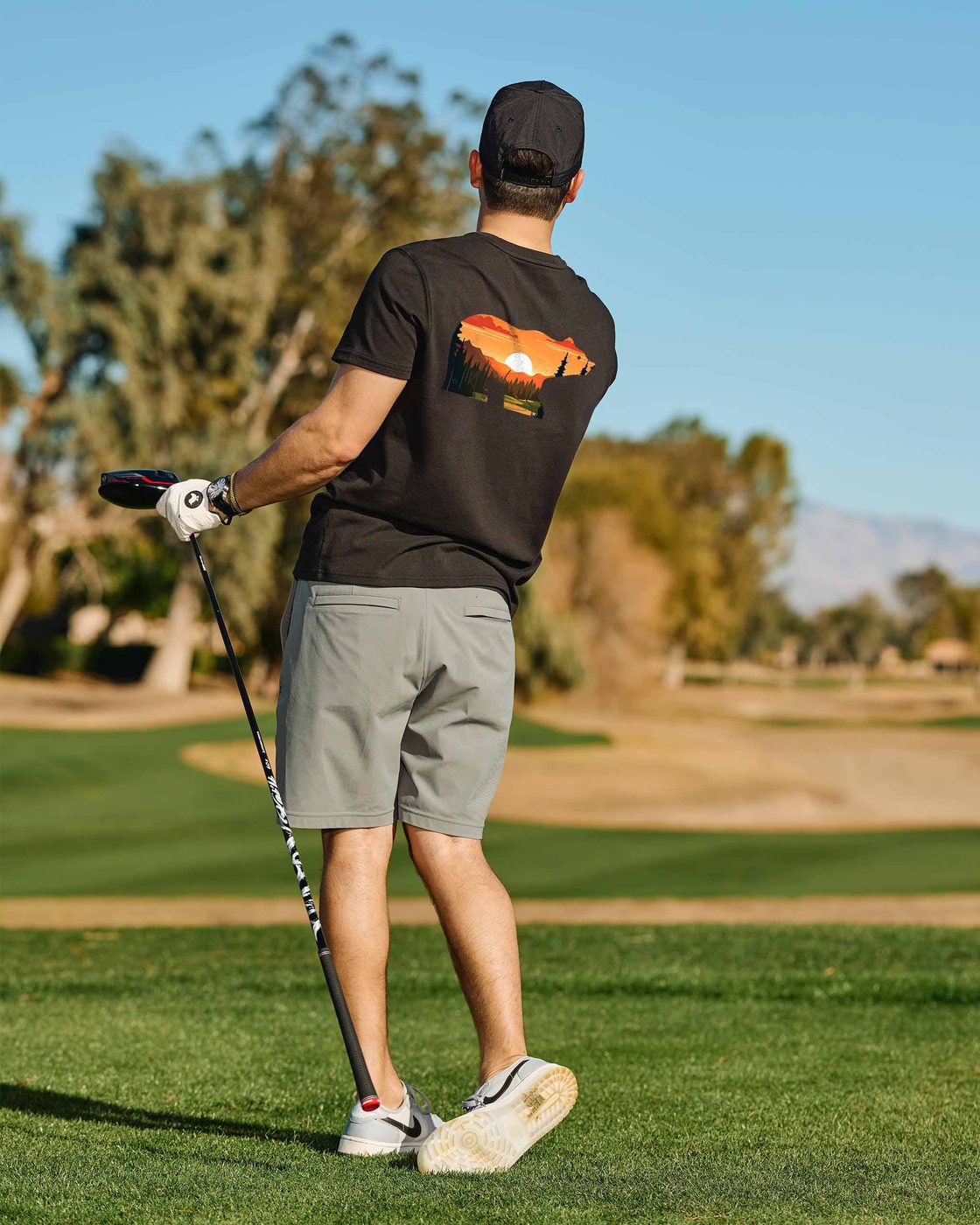 Golfer from behind in a black tee with a large sunset-filled Golden Bear graphic, mid-round on the course