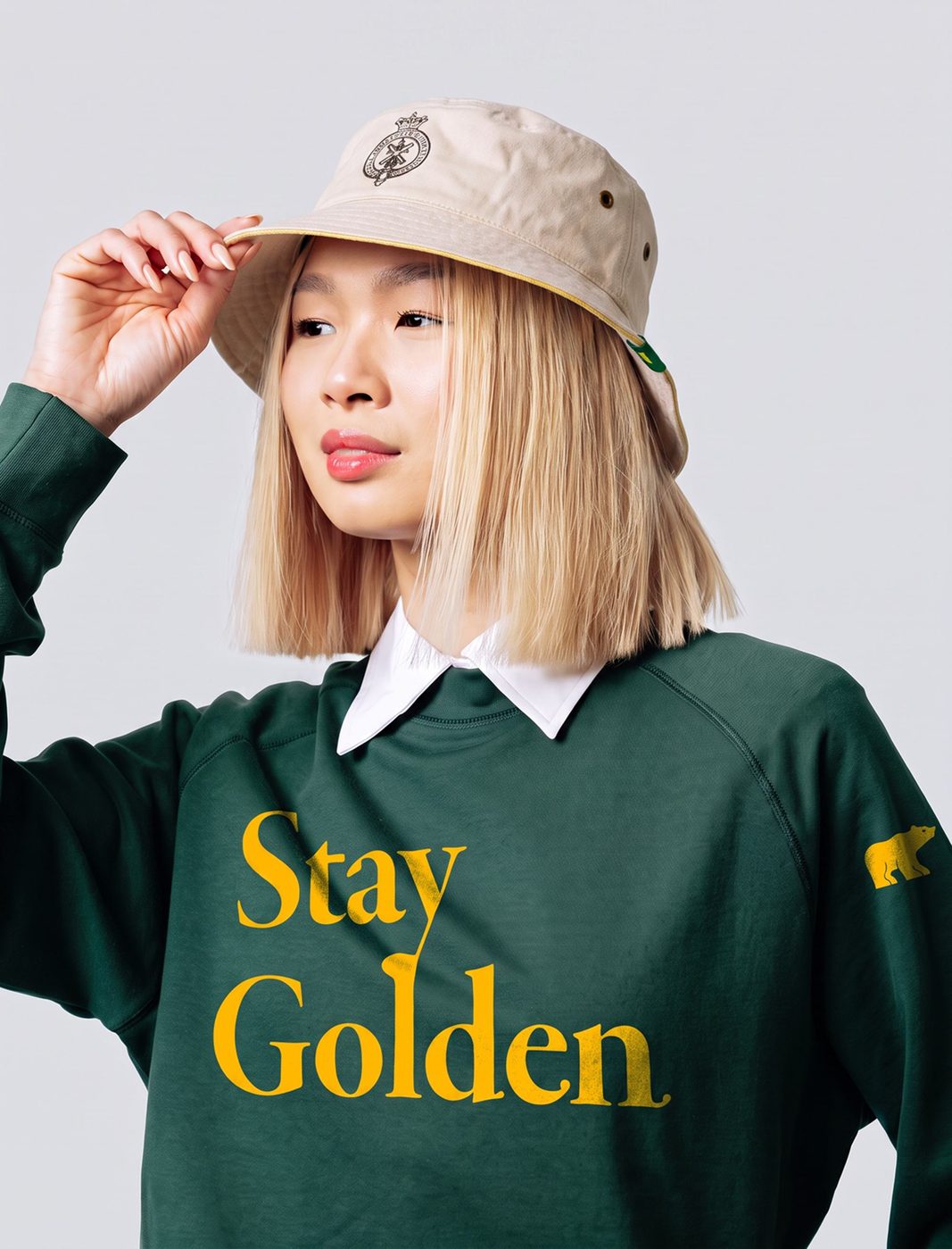 Woman tipping a canvas bucket hat, wearing a forest green Stay Golden crewneck