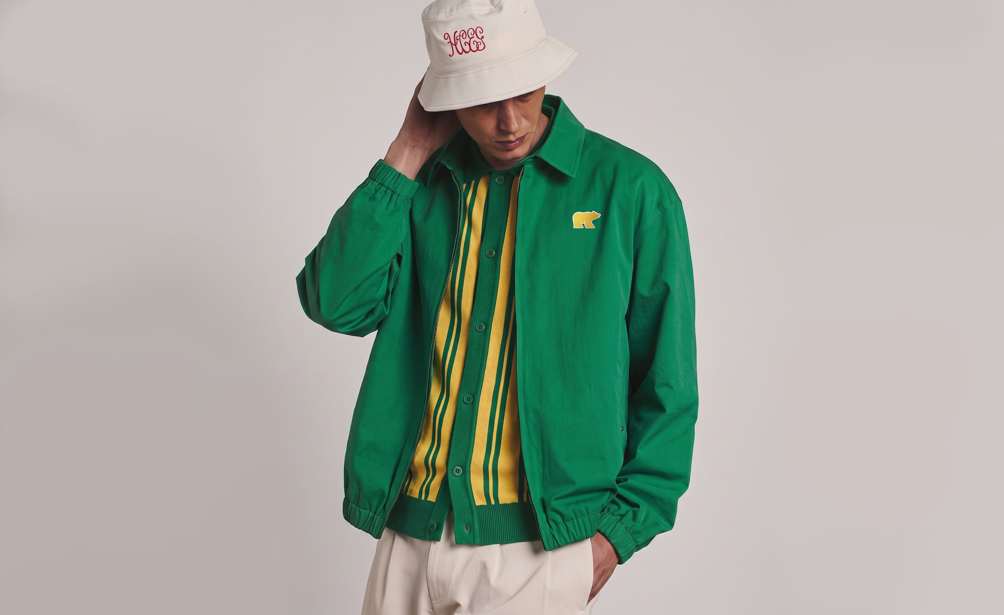 Man in a canvas bucket hat and green jacket with Golden Bear chest patch over a striped knit