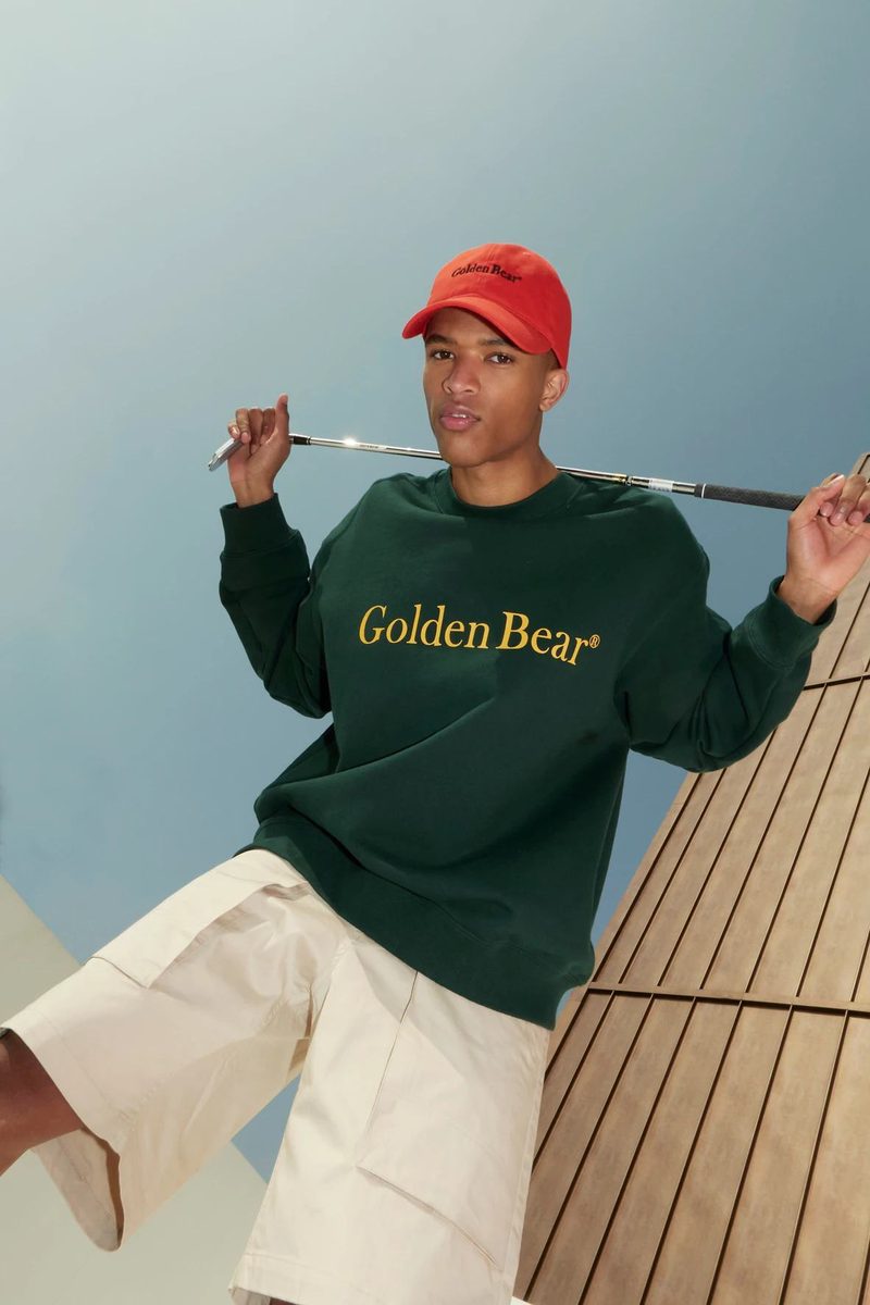 Man in a red canvas cap and forest green Golden Bear crewneck holding a golf club across his shoulders