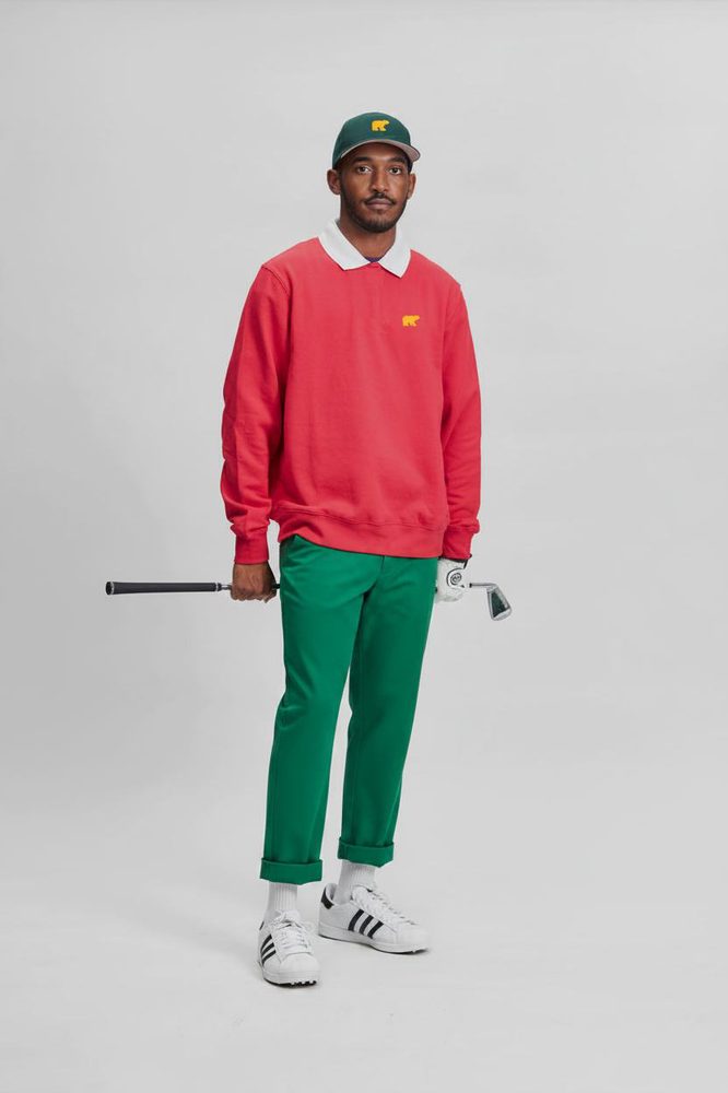 Man in a pink crewneck sweatshirt with white collar, kelly green trousers and a Golden Bear cap