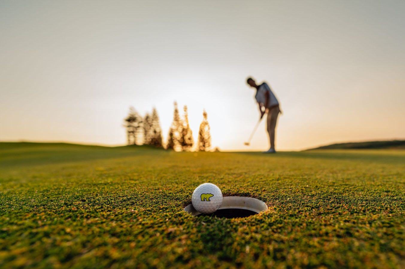 Golfer putting at sunset with a Golden Bear ball resting at the lip of the cup in the foreground