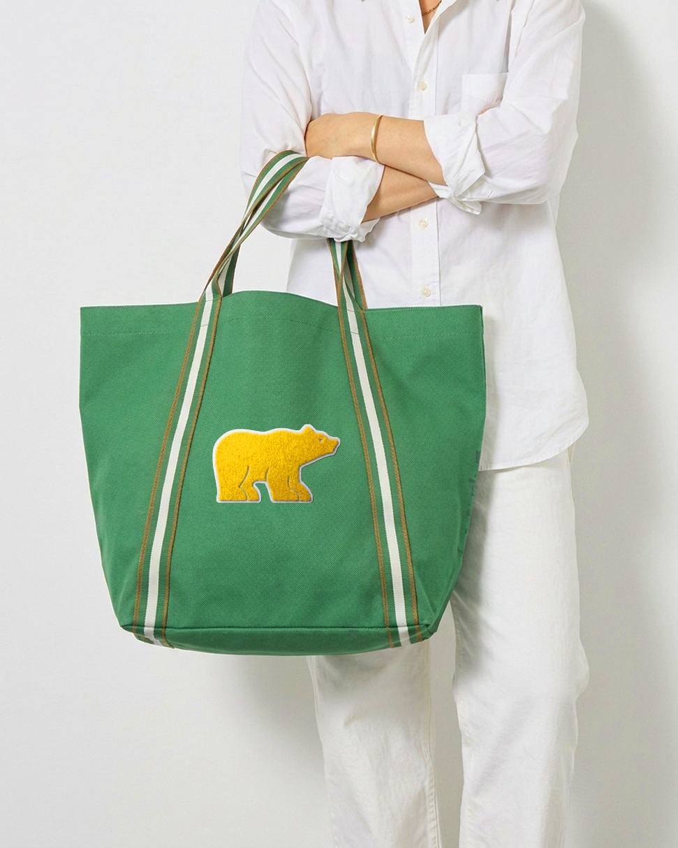 Kelly green canvas tote with a gold Golden Bear chenille patch and striped web handles, carried over a white outfit